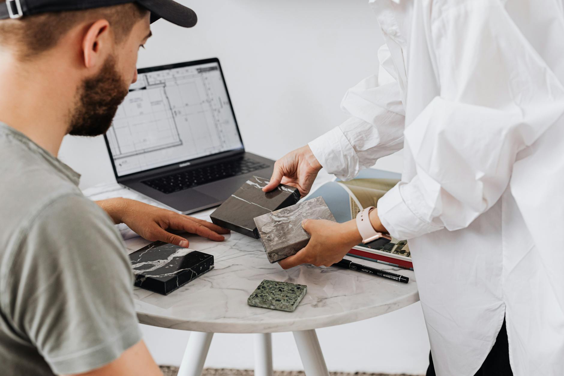 Two designers selecting marble samples indoors with laptop on table.